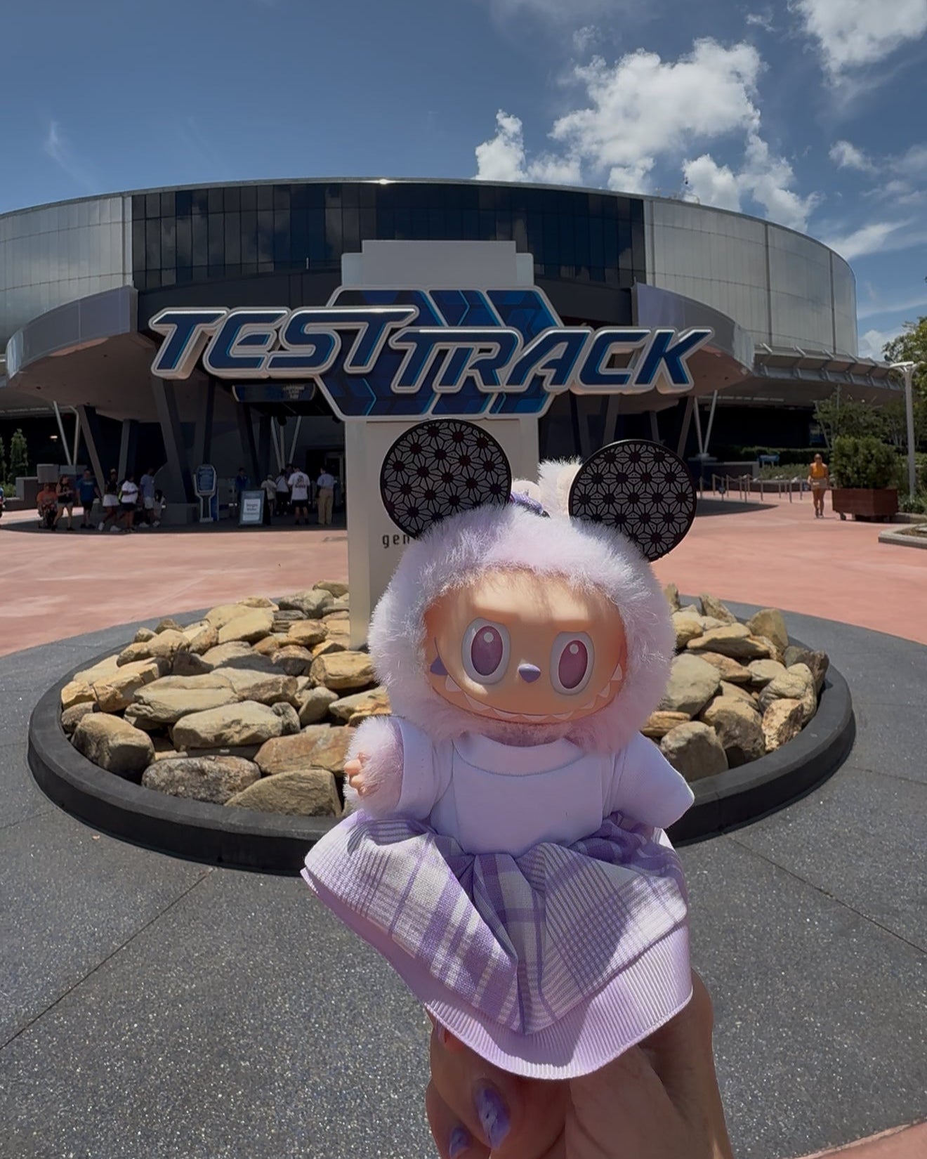 Toy figure with mouse ears in front of a 'Test Track' sign at an amusement park.